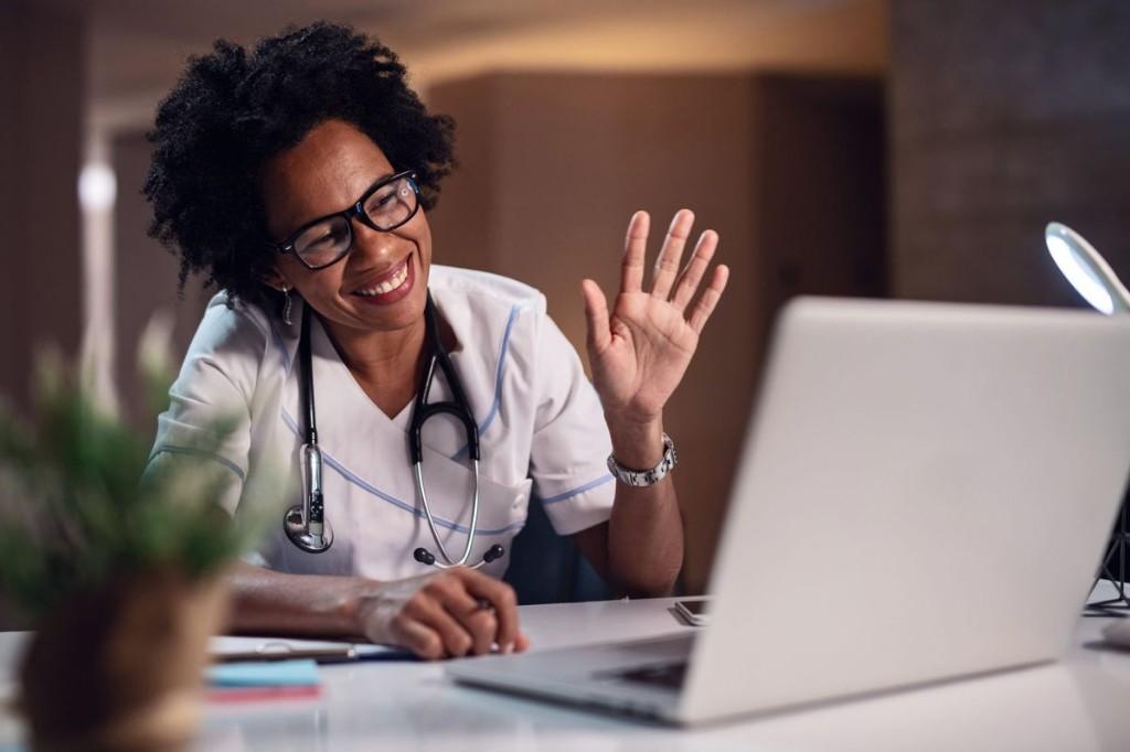 A smiling female doctor in scrubs waving during a video consultation on a laptop, ready to support a patient online